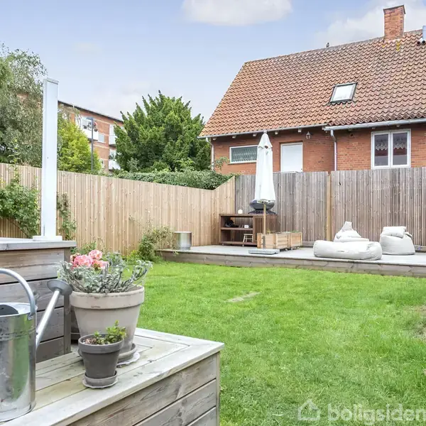 En have med græsplæne, vandkande og blomsterkrukker i forgrunden. I baggrunden ses en terrasse med loungemøbler og en stor parasol foran et rødt murstenshus med et tegltag.