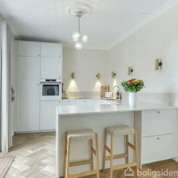 Et moderne køkken med en hvid ø med to barstole, blomster i en vase, og integrerede apparater. Store vinduer til venstre giver lys, og parketgulvet fuldender rummet.