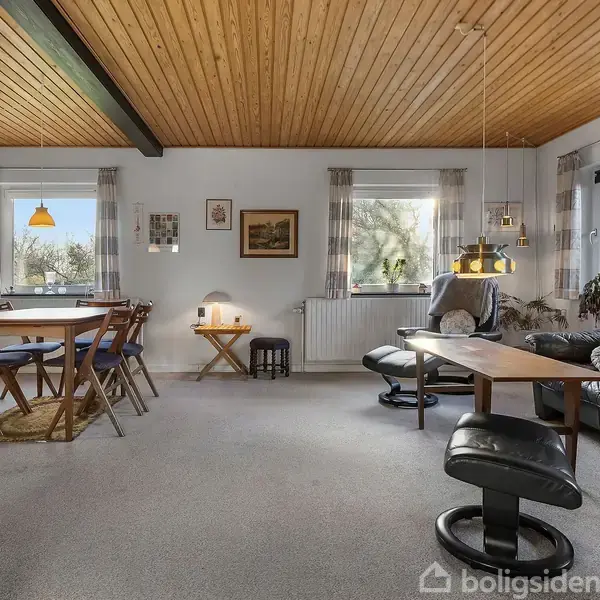 Stue med træloft og gulvtæppe. Sort lædersofa og lænestole omkring et sofabord til højre. Spisebord med stole til venstre. Vinduer med stribede gardiner slipper lys ind.