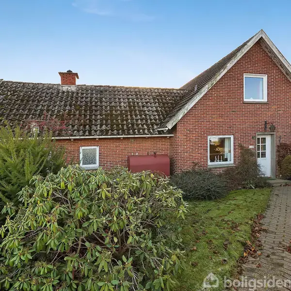Et rødt murstenshus står stille i en have med grønne buske og en flisebelagt sti, under en klar blå himmel.