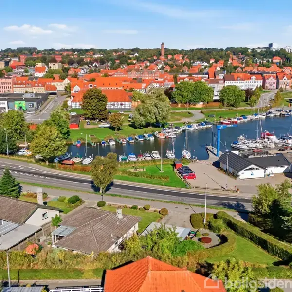 En marina med både ligger ved kanten af en by med røde tegltage og grønne træer. En vej skærer gennem forgrunden, og der ses enkelte huse og bygninger.