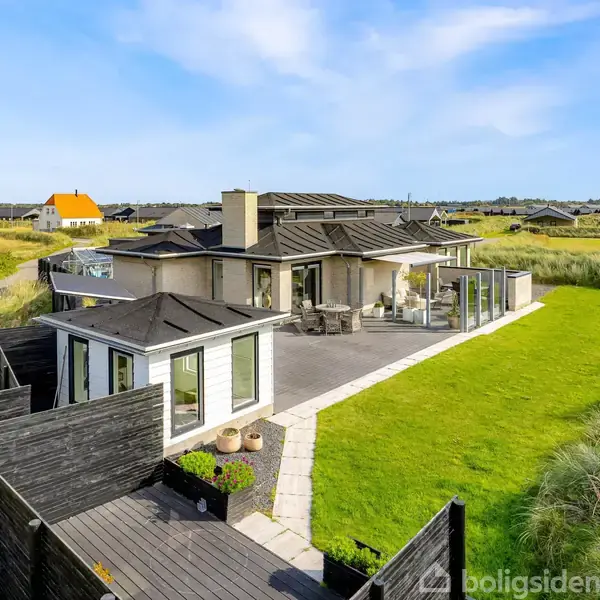 Et moderne sommerhus står på en grøn grund omgivet af klitter. Terrassen har havemøbler og store vinduer med udsigt til landskabet. En sti fører op til hoveddøren.