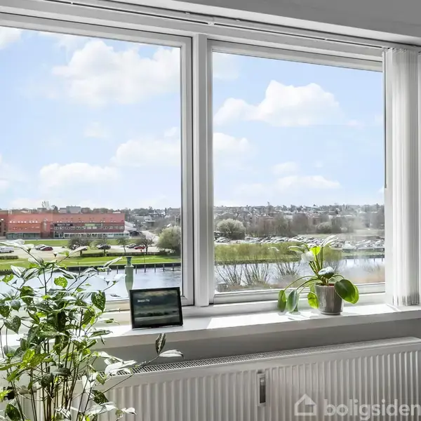 Et stort vindue viser udsigt over en flod med biler og bygninger i baggrunden. Vindueskarmen har en bærbar computer og planter. Sollys belyser rummet med en varm atmosfære.