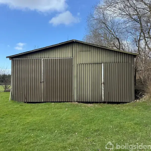 En metalbygning står stille på en grøn græsplæne, omgivet af træer og buskads på en klar dag med blå himmel.