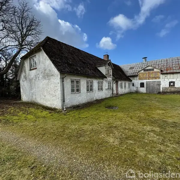 Et gammelt, hvidkalket hus med mørkt tegltag står på en plæne omgivet af træer og en blå skyet himmel. Staldbygningen til højre har en port og træfacade.