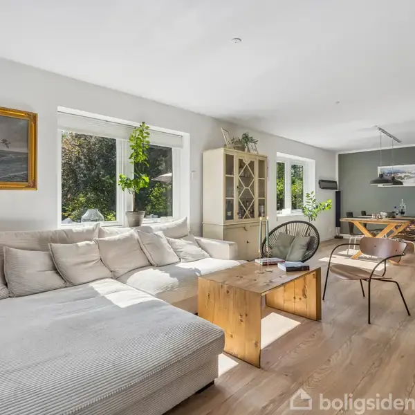 En lys stue med en stor, beige hjørnesofa foran et vindue. Træbord og stole i baggrunden. Grønne planter og et maleri hænger på væggen.