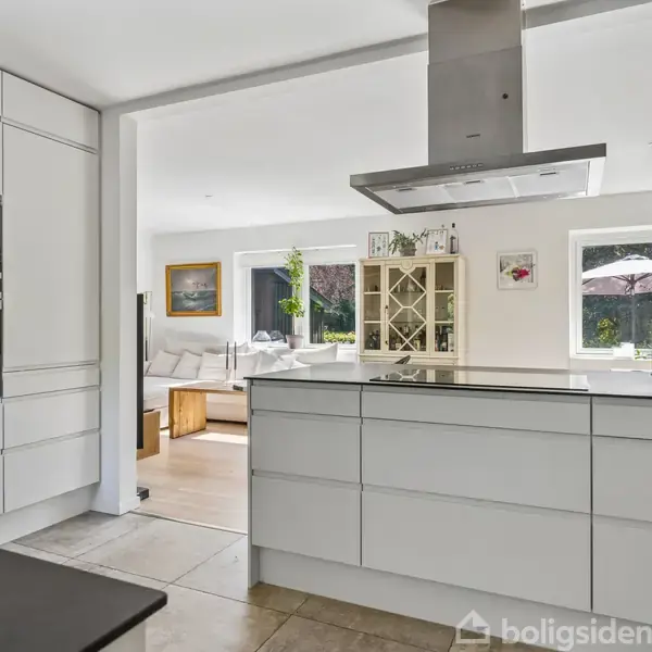 Køkken med moderne, hvidt design dominerer rummet. Kogeøen har en emhætte over sig. I baggrunden er en lys stue med sofa og reoler, forbundet til køkkenet.