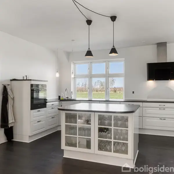 Køkken med en central ø med glaslåger og sort bordplade, hængende lamper, hvide skabe og en ovn. Store vinduer giver udsigt til en grøn have. Ingen tekst til stede.
