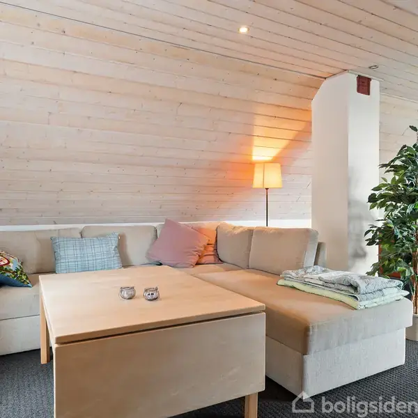 En beige sofa står i en stue med skrå loft. Et sofabord er foran, med lys og puder ovenpå. En gulvlampe lyser i hjørnet, ved siden af en stor grøn plante. På gulvet ligger en mørkeblå tæppe.
