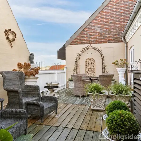 Rattanmøbler står på en træterrasse omgivet af plantekasser og vægdekorationer i en hyggelig gårdhave. En mur med dekorativ bue og væghegn skaber et indbydende miljø.