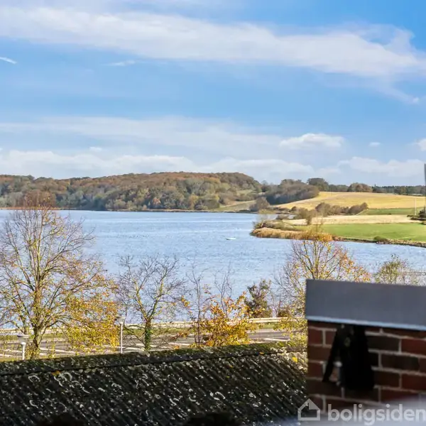 Udsigt over en sø med træer i efterårsfarver langs bredden. I baggrunden ses bakker og åbne marker under en blå himmel med lette skyer.