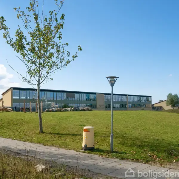 En moderne bygning står stille på en grøn græsplæne med spredte træer og en brolagt sti i forgrunden. En lygtepæl og skraldespand er synlige.