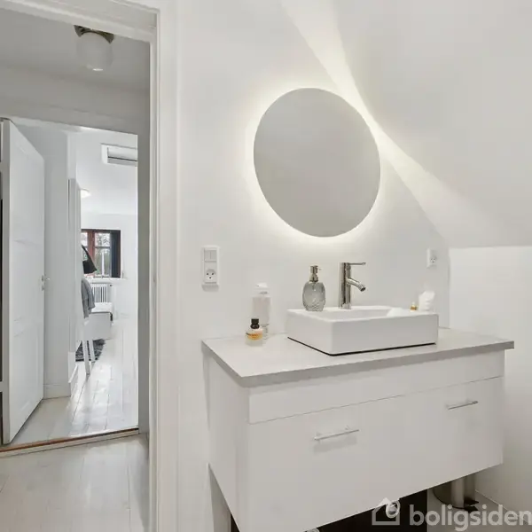 Rundt spejl med lys hænger over en vask med armatur på hvidt skab. Håndklæde hænger på væggen. Døråbning fører til et lyst rum i baggrunden.