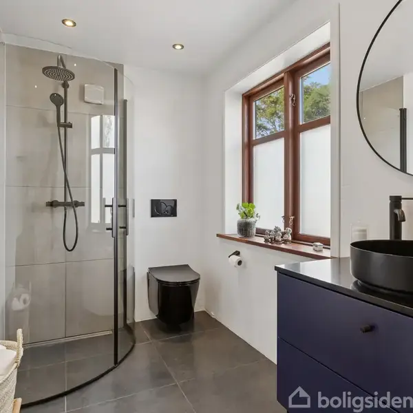 Bruser med glasvæg står i et moderne badeværelse. Ved siden af findes et sort toilet og en sort vask på et mørkt skab. Et stort vindue lader naturligt lys ind.