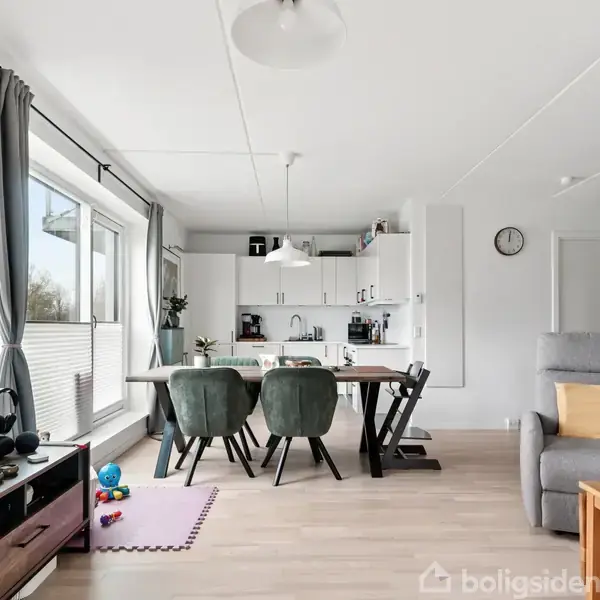 Stue-køkken med spisebord og stole, åbne hylder med pyntegenstande, en stor fladskærm på væggen, og farverige legetøj på gulvet ved vinduer, der slipper dagslys ind.