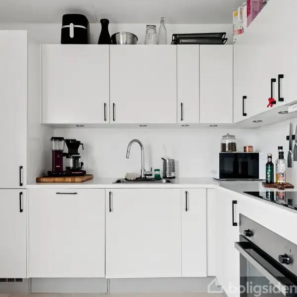 Hvidt køkken med hvide skabe og sort håndtag, hvor en kaffemaskine står ved vasken. Flere køkkenredskaber hænger på væggen, og hvem ved siden af en ovn og kogeplade.