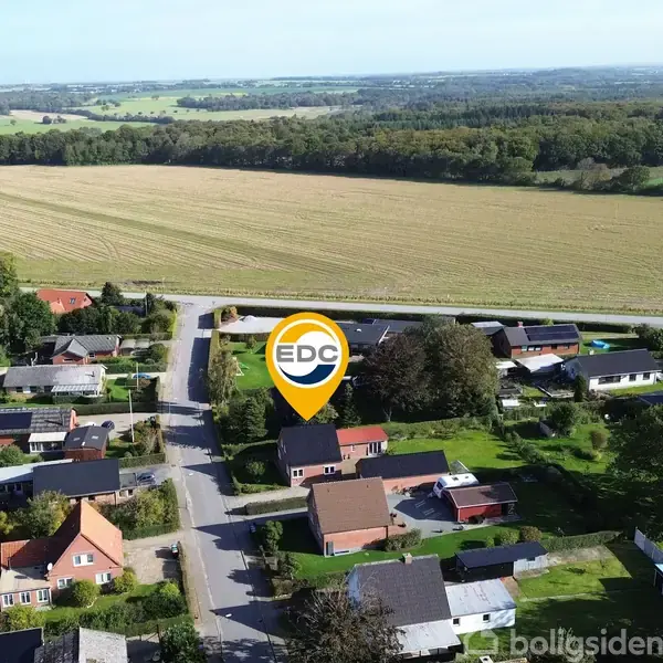 En bolig markeret med et "EDC"-ikon står centralt i et villakvarter. Omkringliggende huse er omkranset af marker og skov i det fjerne.