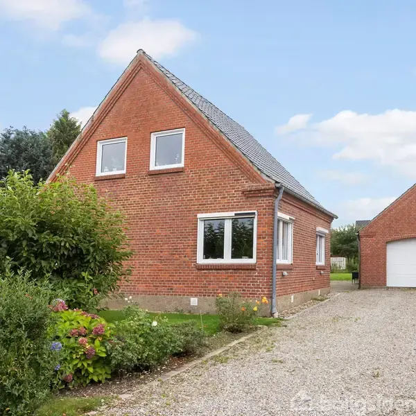 En rød murstensvilla med kvist står roligt i en have omgivet af grønne buske og blomster, en grusvej fører til en separat garage i baggrunden.