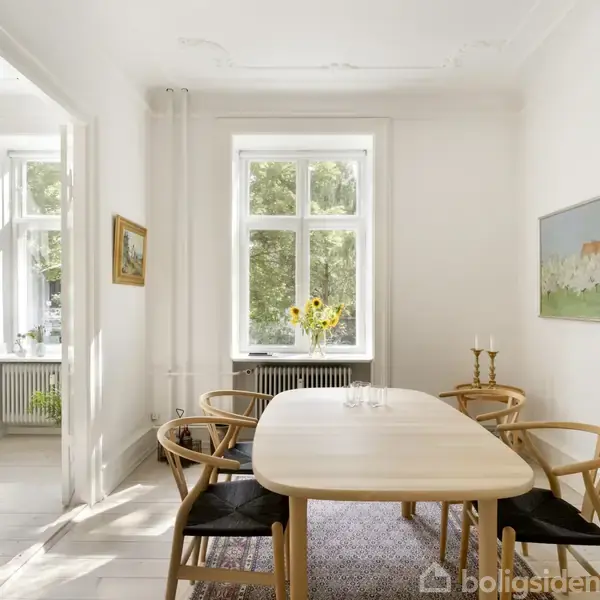 Et lyst spisebord er omgivet af seks stole i en hvidt malet spisestue. Et vindue med udsigt til træer bagved indeholder solrige blomster. To billeder dekorerer væggen.