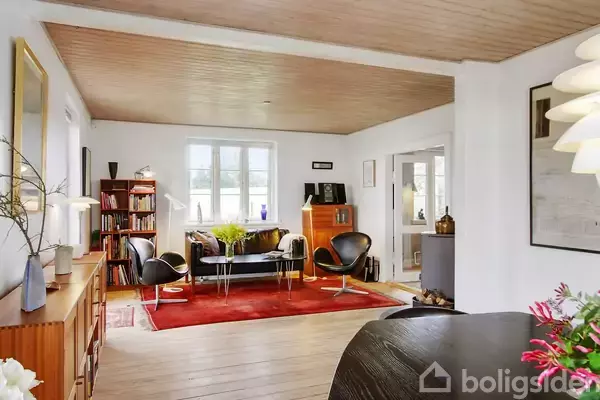 Stue med møblering: sorte stole og sofabord på rød tæppe, i lys og velindrettet rum med træloft og vinduer. Omgivet af bogreoler og dekorative elementer, klassisk indretning.