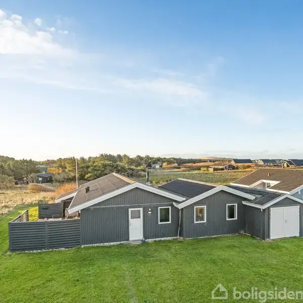 Hus med gråt træbeklædning og hvide vinduer står stille i en grøn have. Omgivet af et naturskønt landskab med flere huse i baggrunden under en blå himmel.