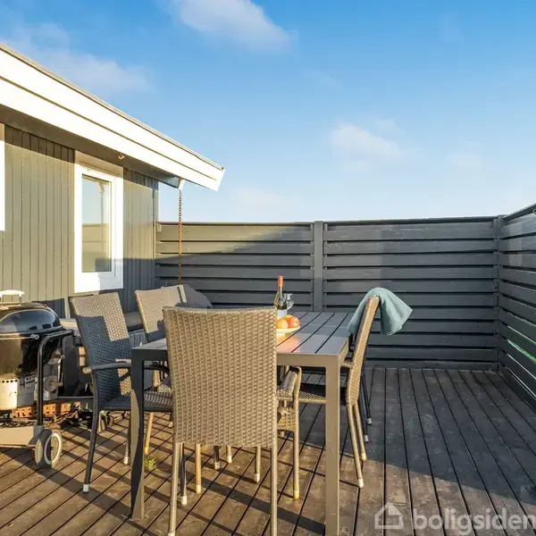 En terrasse med en bordgruppe på seks stole og en kulgrill. En flaske vin og et glas står på bordet. Omgivet af højt hegn og en husvæg.