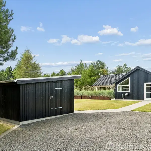 Et sort hus og skur står i en velplejet have med græsplæner og buske. Stier af småsten forbinder bygningerne, og skov omkranser området under en blå himmel.