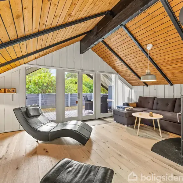Et moderne loftsrum med skrå trælofter og store vinduer. Der står en sort loungestol ved siden af en pejs, mens en sofa og et bord danner siddeområde. En terrasse ses udenfor.