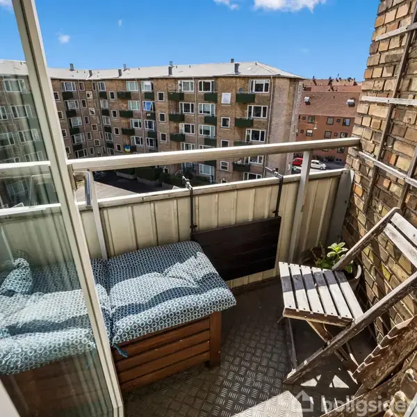 En lille altan med blåmønstrede hynder på en bænk ved siden af en træstol. Altanen har udsigt til et boligkompleks i baggrunden under en blå himmel.