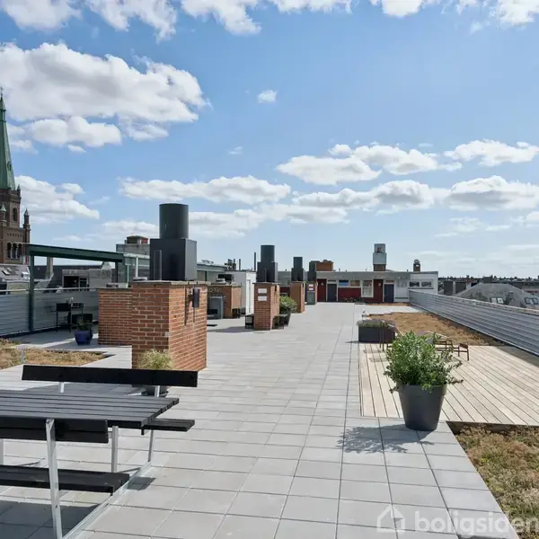 Tagterrasse med borde og bænke, planter i potter, og byudsigt. En kirketårn ses i baggrunden under en blå himmel med spredte skyer.