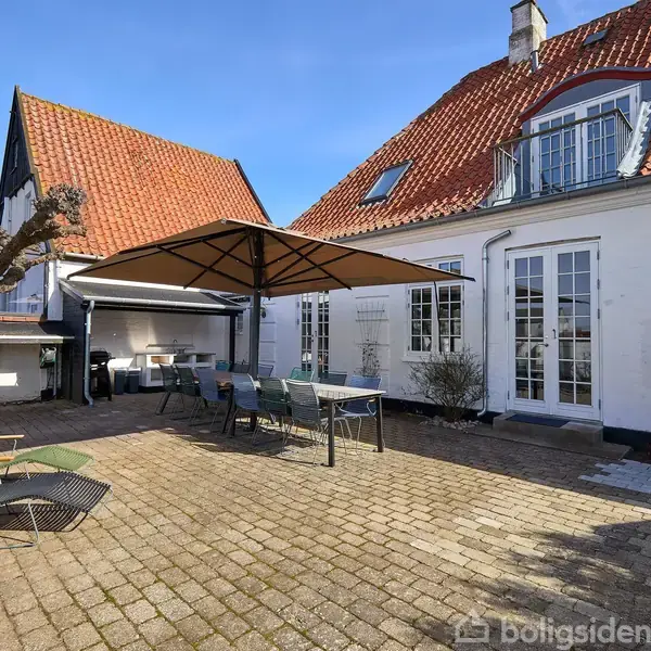 Et gårdhaveområde med et stort bord under en parasol. Omkring står flere stole. Til venstre ses et prydtræ og to liggestole. Huset har rødt tegltag og hvide mure.