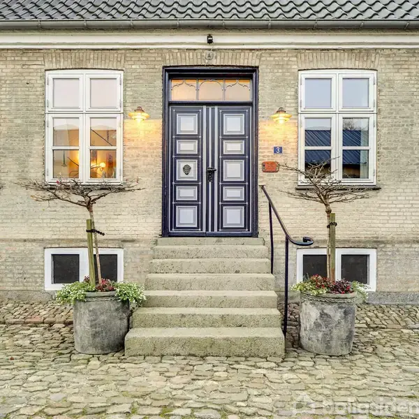 Dør med trapper foran et murstenshus. To planter står i potter på hver side af trappen. Vinduer flankerer døren. Husets facade er flisebelagt og omgivet af stenvægge.
