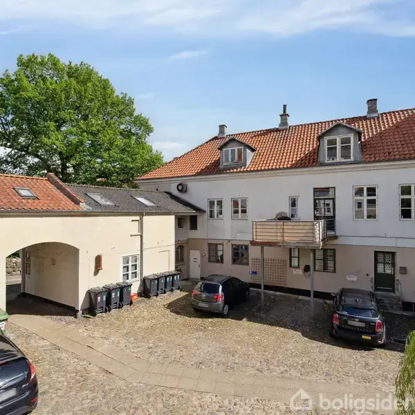 Gårdhave med to parkerede biler foran en hvid bygning med rødt tegltag. Bygningerne omkranser en brolagt plads. En stor grøn træ ses i baggrunden.