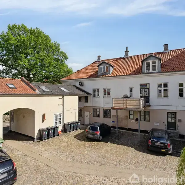Flere biler holder parkeret på en brostensbelagt gårdsplads foran en hvid toetagers bygning med rødt tegltag. Grønne træer står i baggrunden under en blå himmel.