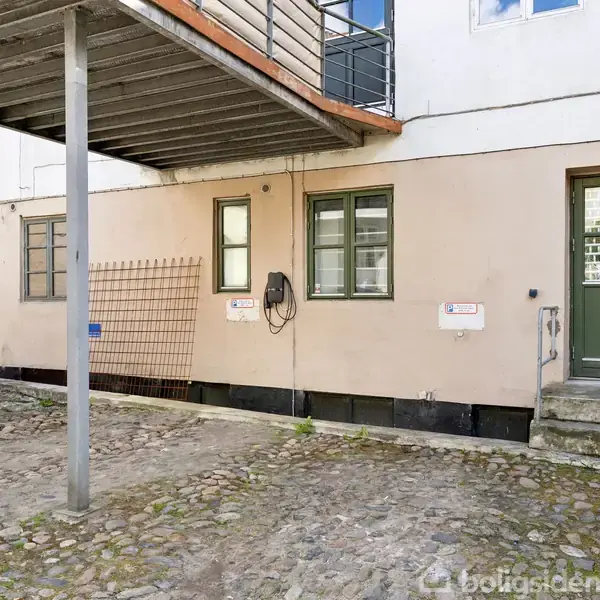 Billede af en baggård med brosten. En carport ses til venstre, og en hoveddør op ad et par trin til højre. To små vinduer ses på væggen.