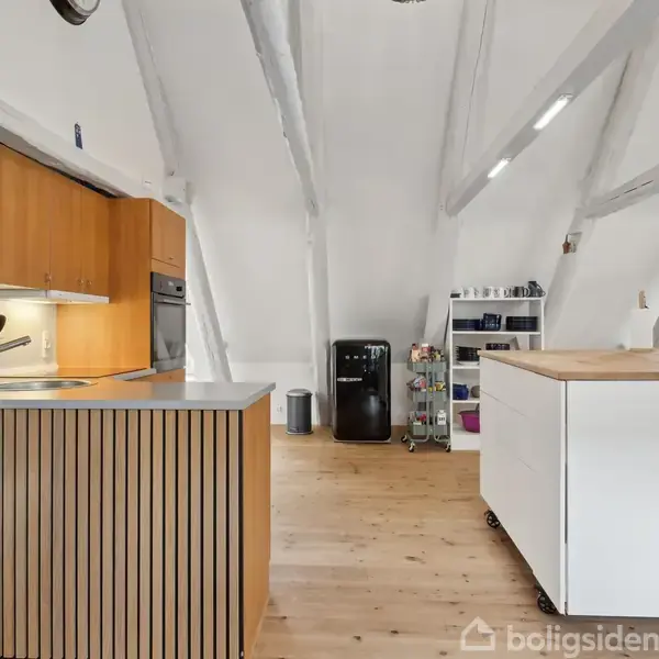 Et moderne køkken med træelementer fylder et rum med skrå lofter. En køkkenø står centralt, mens et køleskab og hylder ses i baggrunden. Væggene er hvide.