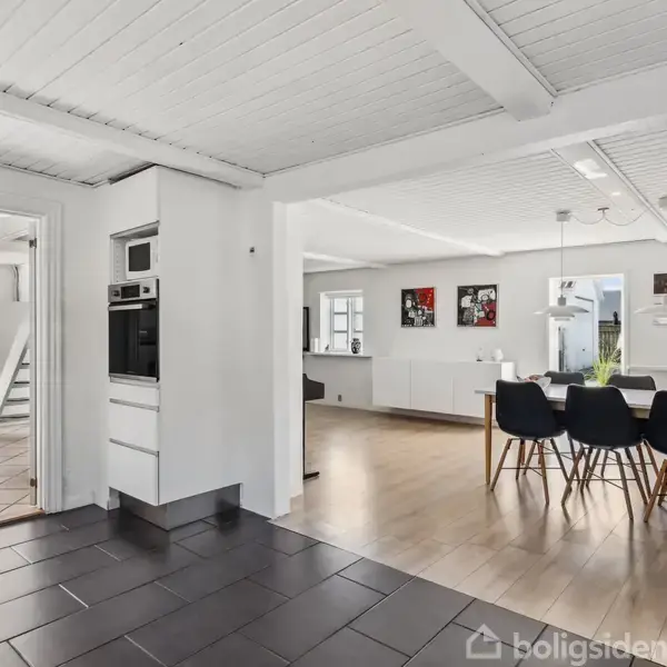 Et moderne køkken-alrum med spisebord og sorte stole står midt i et lyst rum med trægulv, hvide vægge og synlige bjælker. Bagved ses en dør og en trappe.