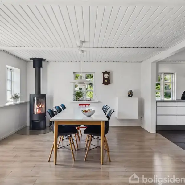 En spisebord med fire stole står i et lyst rum med trægulv. En brændeovn opvarmer rummet ved siden af et åbent køkken med hvidt interiør.