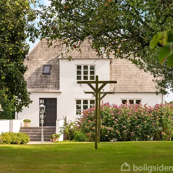 Et hvidt hus står i en frodig have med grøn græsplæne. Omkring huset er der træer og blomster. Kreditupload er "Boligsiden".