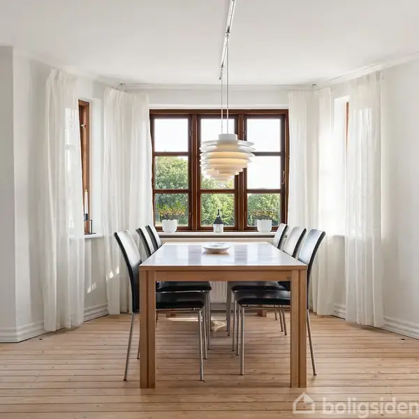 Et rektangulært spisebord med seks sorte stole står i et lyst rum med trægulv. En moderne lampe hænger over bordet, og vinduerne bagved giver udsigt til grønne træer.