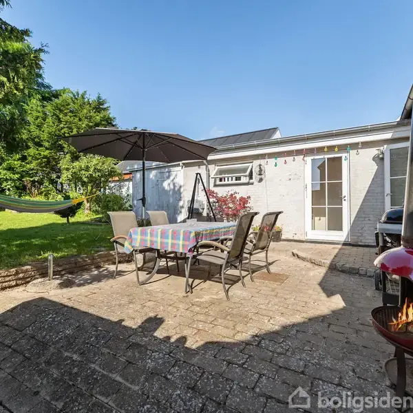 Terrasse med spisebord dækket af stribet dug og stole under parasol, omgivet af græsplæne og træer. En rød udendørs pejs står til højre, og en hængekøje er til venstre.
