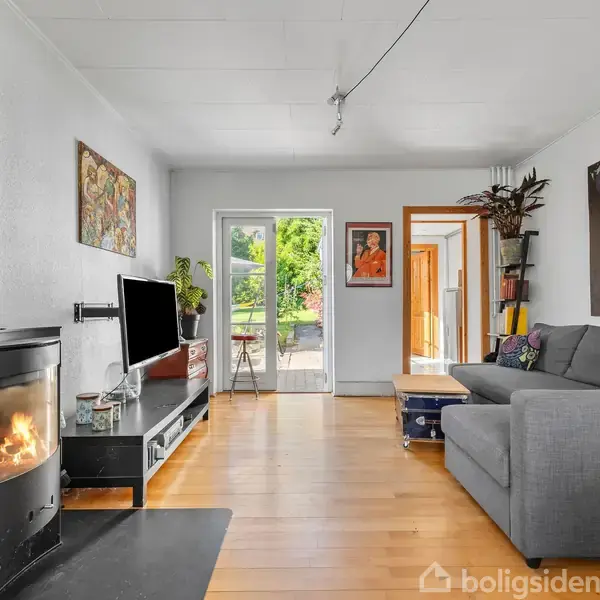 Brændeovn med ild står i stue ved siden af sort tv-møbel. Grå sofa og kunst på væggen. Glasdøre fører ud til en terrasse med grønt landskab.