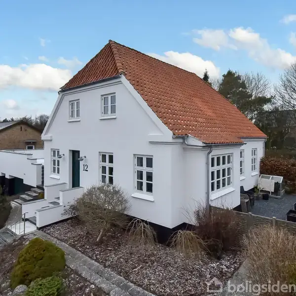 Et hvidt hus med rødt tag står i en have med buske og stier. Tallet "12" vises ved døren i en fredelig, forstadsagtig omgivelser.