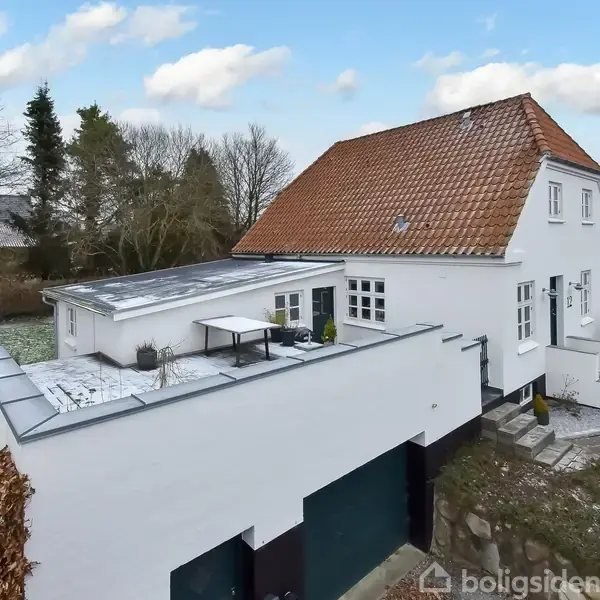 En hvid to-etagers villa med rødt tegltag står i en snoet have med træer. Terrassen på taget har et bord og stole. Snepletter ses på jorden.