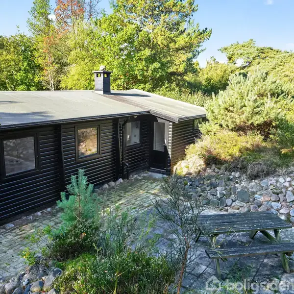 Træhus med sorte vægge står mellem træer i en have. Foran huset er en stenbelagt terrasse med et træbord og bænke. Sollys filtrerer gennem træerne.