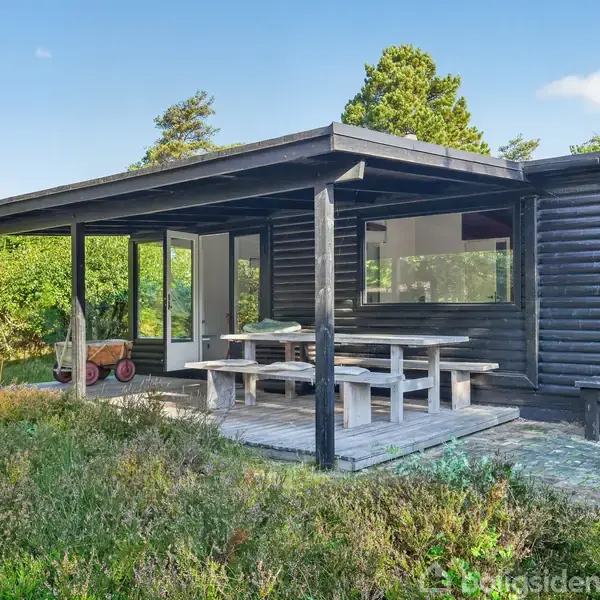 Træsommerhus står på en terrasse med bordbænkesæt, omgivet af frodige buske og træer under en blå himmel.