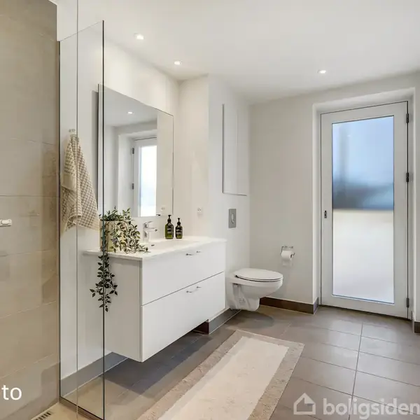 Et moderne badeværelse med en brusekabine til venstre, en håndvask med spejl foran, og et toilet ved siden af. Lyse fliser og en dør leder udenfor. Tekst: "Modelfoto".