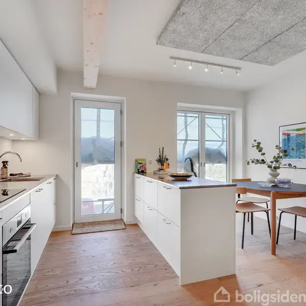 Et moderne køkken med hvidt inventar og en ø i midten. Til højre står et spisebord med to stole og vægkunst. Store vinduer giver naturligt lysindfald. Tekst: "Model foto"