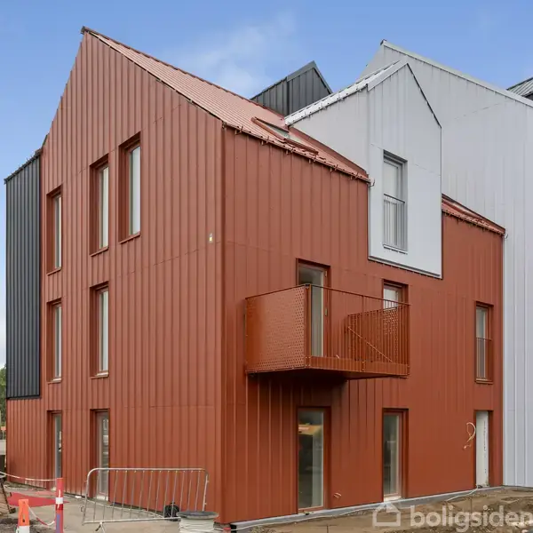 En moderne etagebygning har røde, sorte og hvide facader, omgivet af en byggeplads med byggehegn og materialer. Bygningen har store vinduer og en lille balkon.