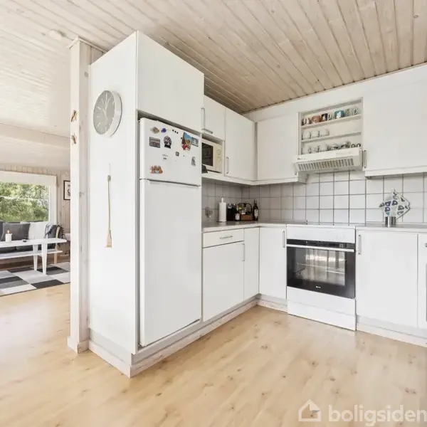 Hvidt køkken med hvidevarer, trælaminatgulv, og flisevæg. Køleskabet har magneter, og der er en vægmonteret ur. Åben stue i baggrunden med hvide møbler og store vinduer.